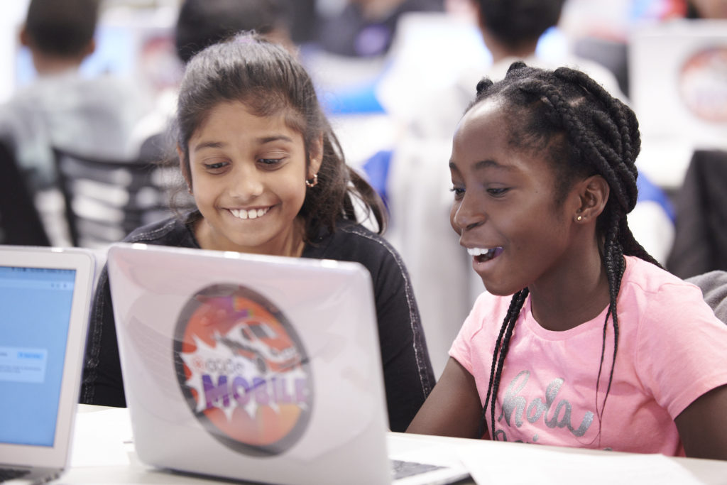 Two girls are sitting  and smiling in front of laptop