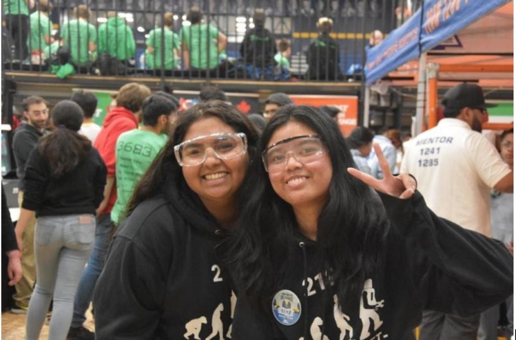 Two female-identified teens are wearing lab glasses  and smiling