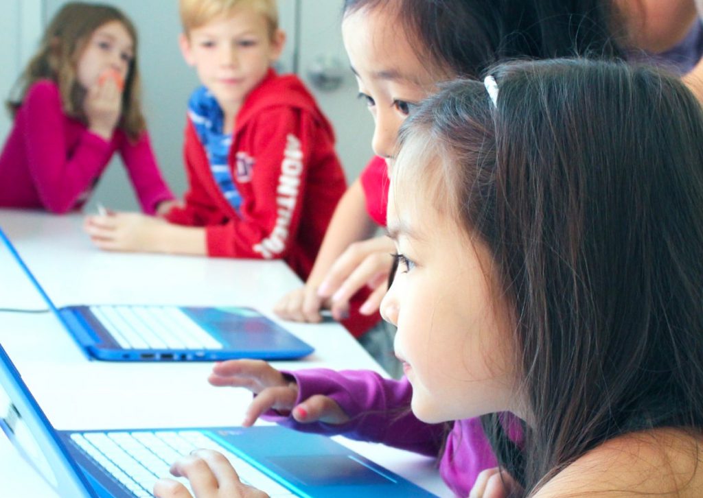 two girls looking working on a laptop with children in the back ground