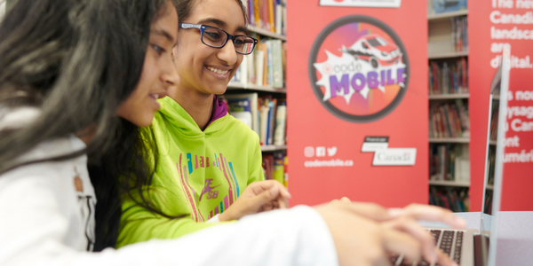 two girls on their laptops in a library and the Code Mobile Logo in the back